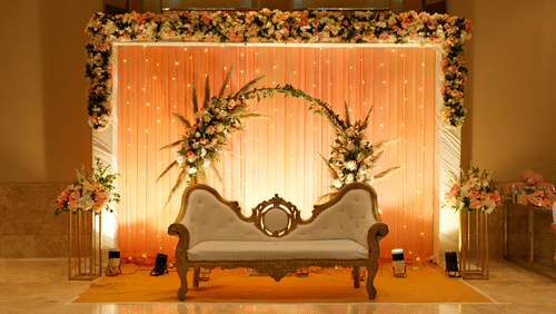 A wedding stage with a floral backdrop, seating for two, and elegant lighting.