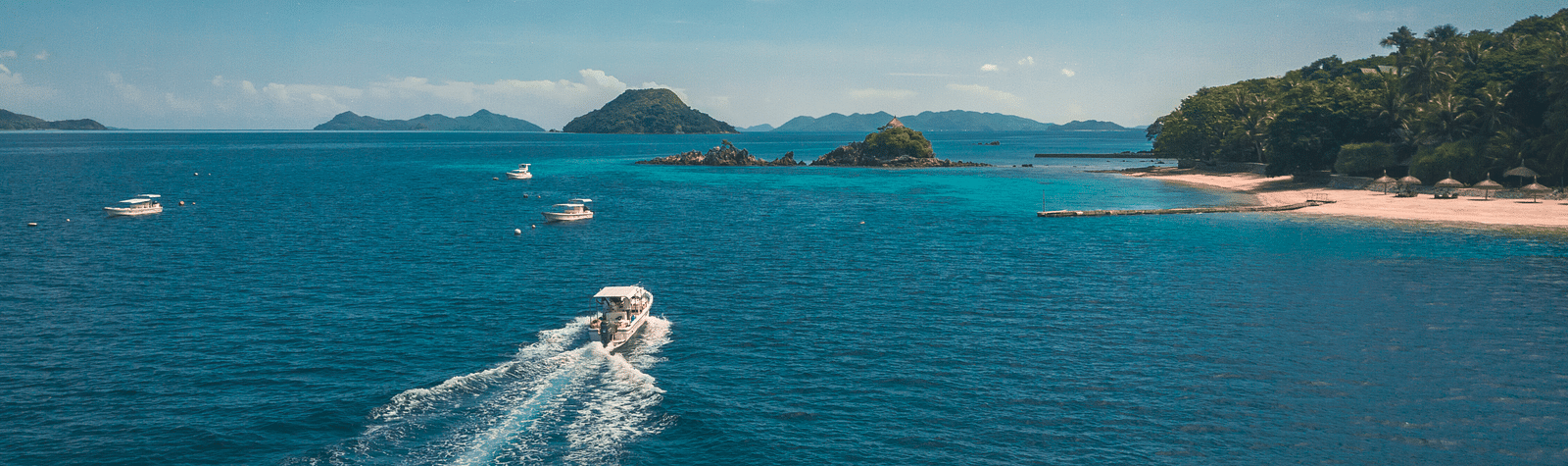 Aerial view of a speedboat cruising through turquoise waters near a tropical island, with white sandy beaches, lush greenery, and distant mountain isles under a clear blue sky.