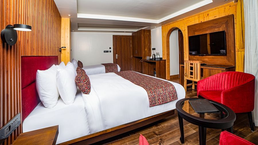 Bright and inviting hotel room with a large bed featuring red accents, warm wood panelling, a flat-screen TV, a wooden work desk, and a cosy seating area with red armchairs and a round glass-top table - Yashshree Lachen.