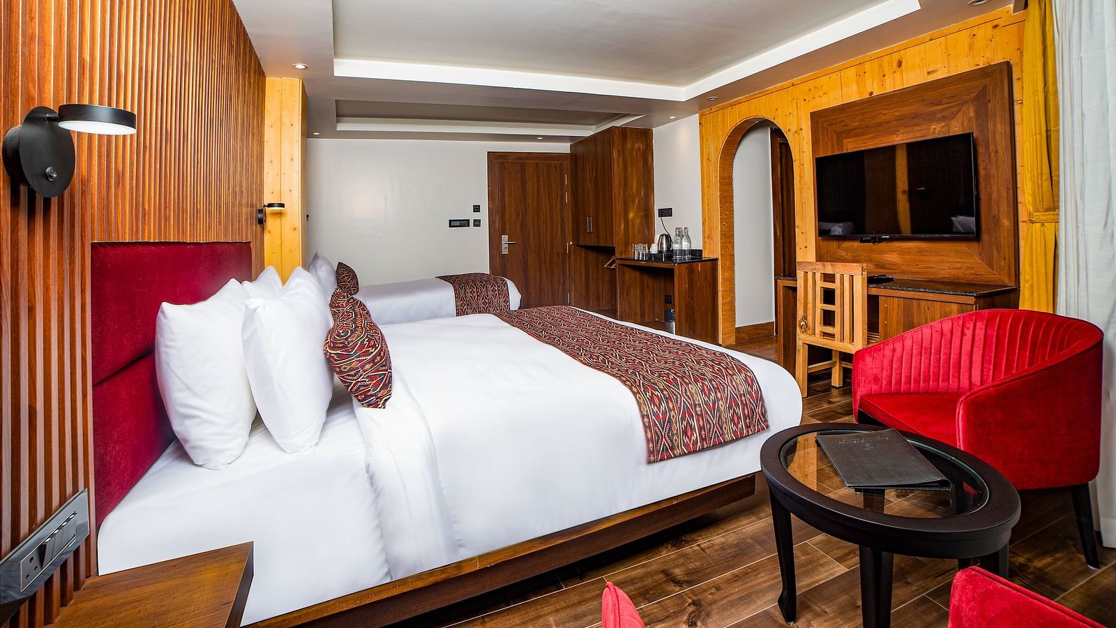 Bright and inviting hotel room with a large bed featuring red accents, warm wood panelling, a flat-screen TV, a wooden work desk, and a cosy seating area with red armchairs and a round glass-top table - Yashshree Lachen.