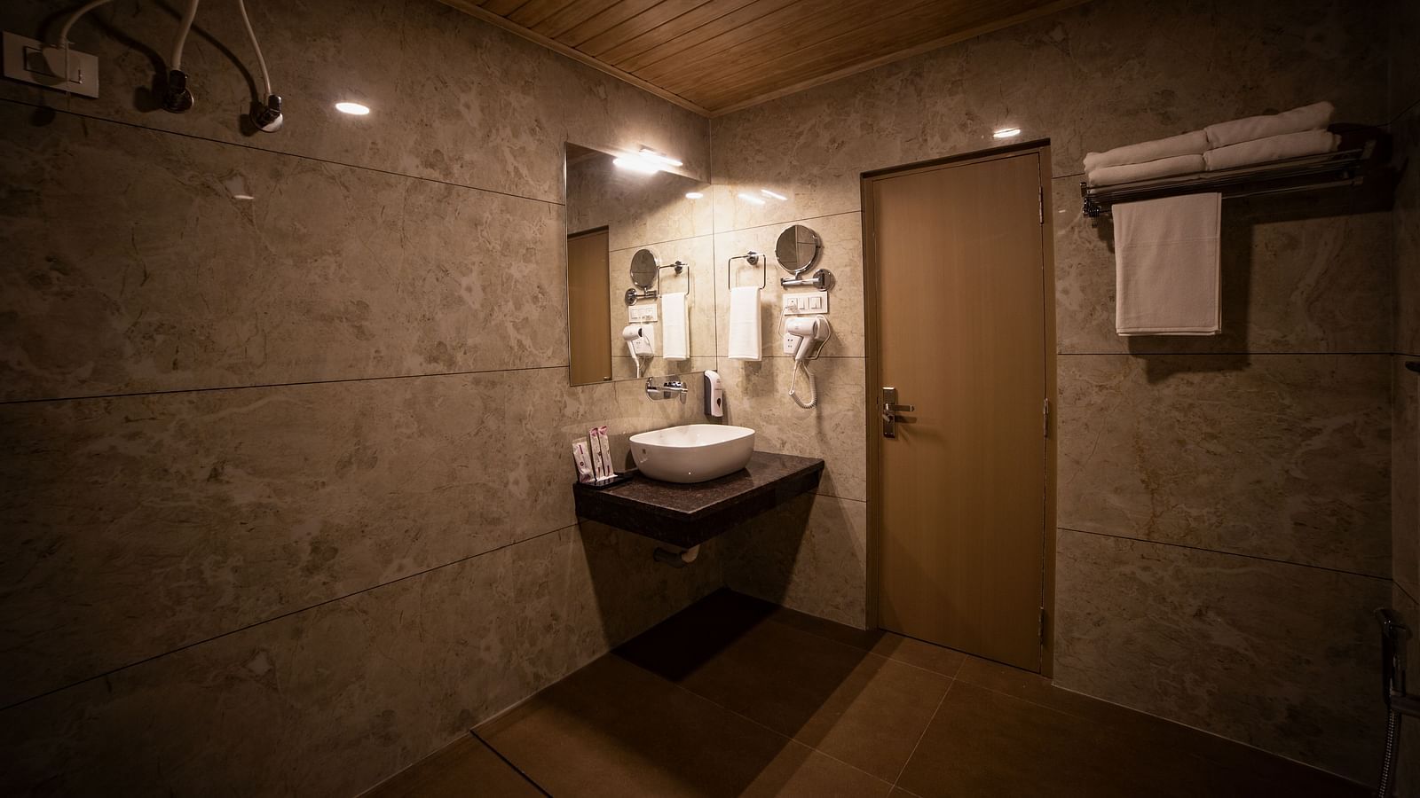 Modern bathroom with beige marble walls, a floating sink with a large mirror, a wooden door, towel racks, and soft ambient lighting - Yashshree Pelling Chekhim Retreat.