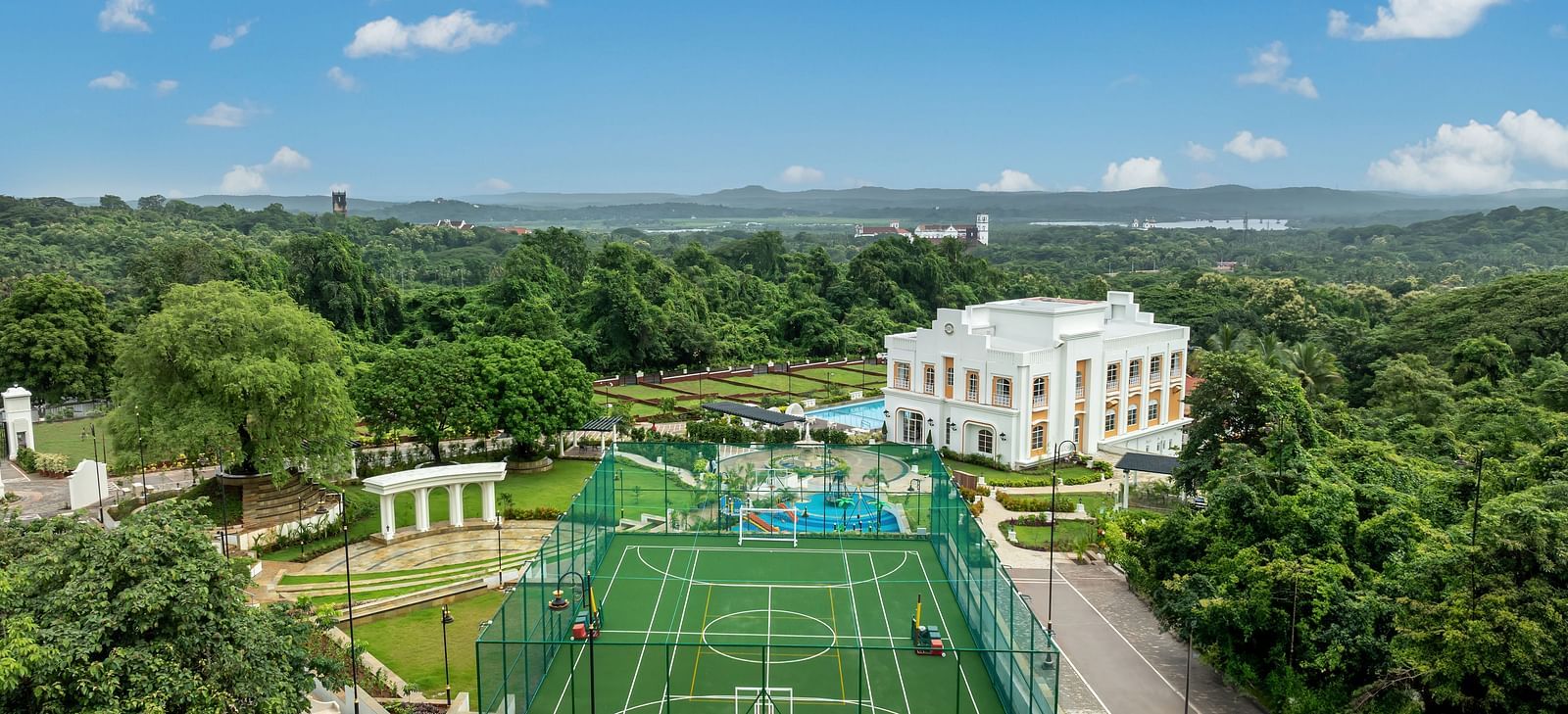 A drone shot of Imperial Goa featuring a multi-purpose ground along with the bright white coloured villa surrounded by lush greenery.