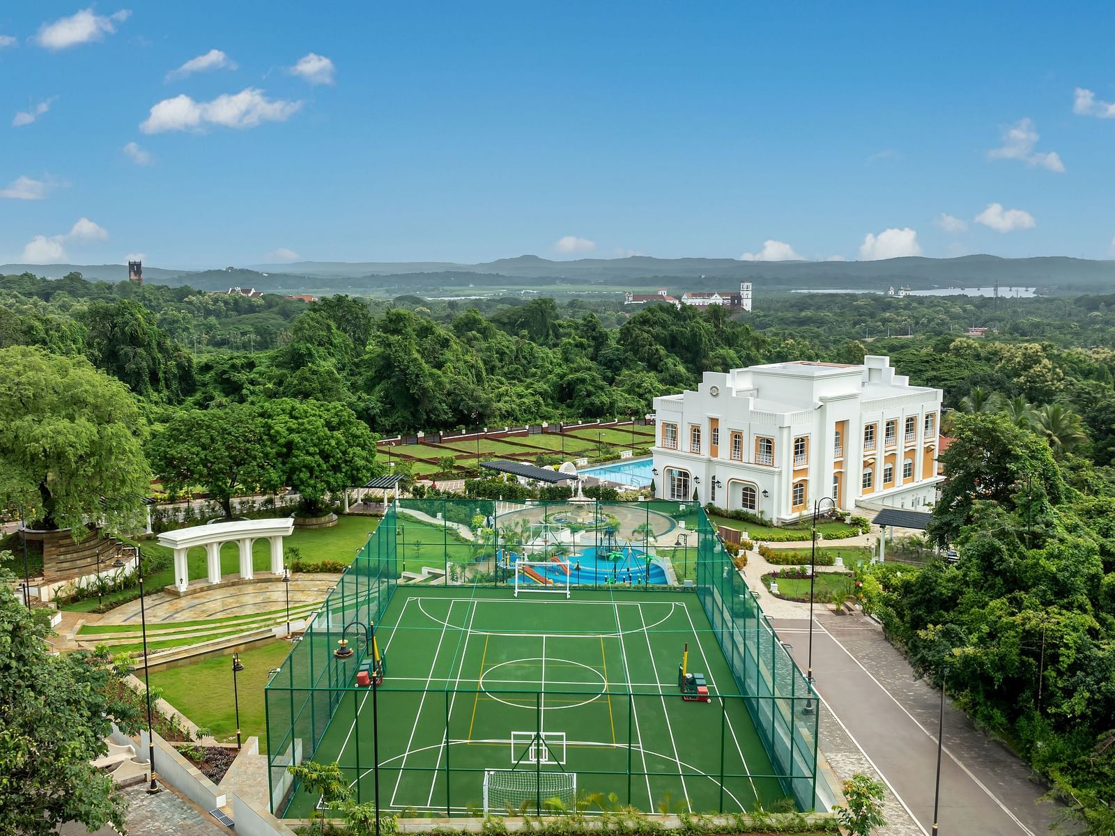 A drone shot of Imperial Goa featuring a multi-purpose ground along with the bright white coloured villa surrounded by lush greenery.