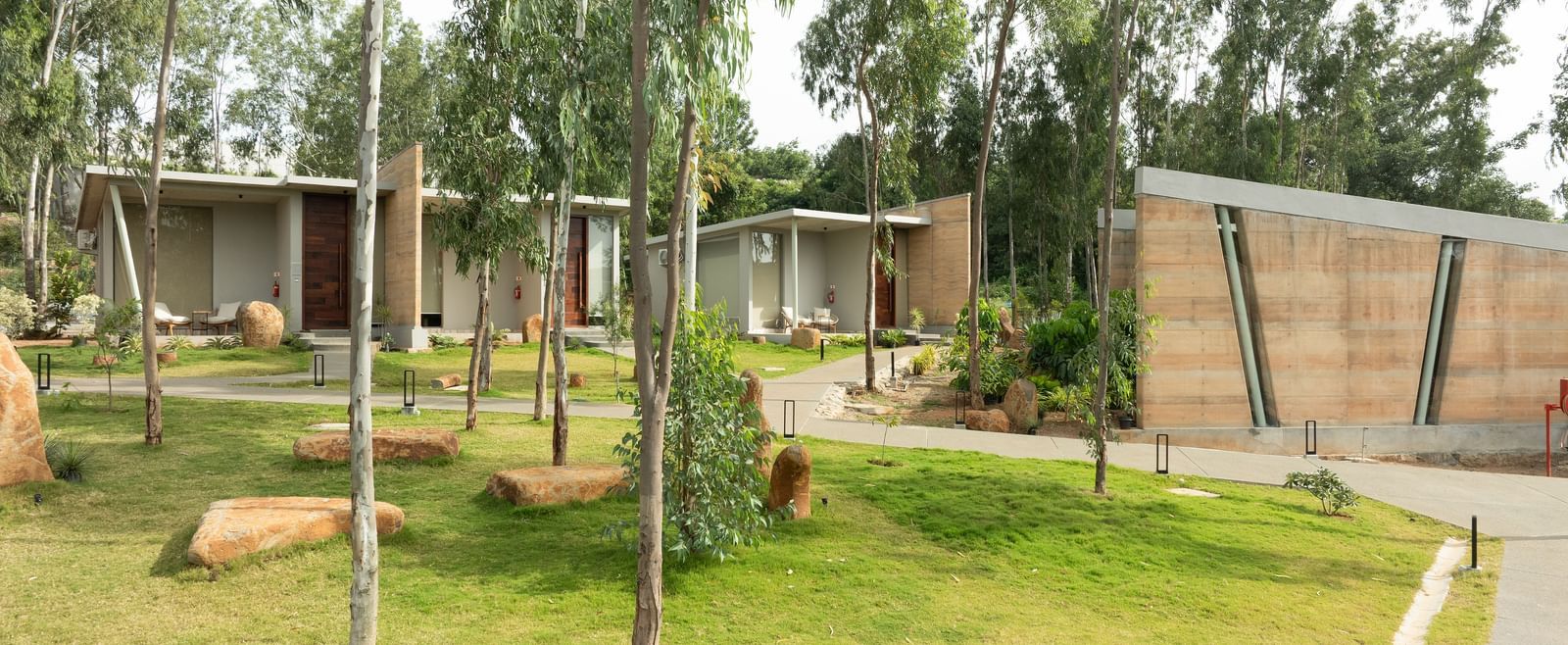 A wide exterior shot showing multiple modern residential units nestled within a landscaped area with trees and pathways | Gaia Studio | RD's Nature Retreat, Bangalore