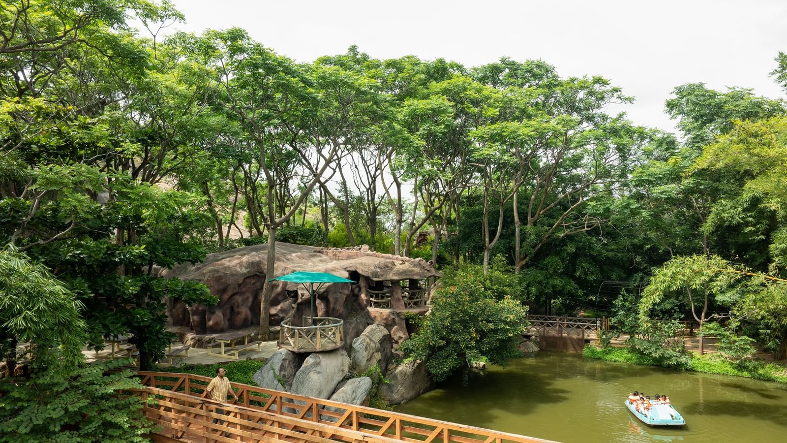 A tranquil pond with a wooden walkway leads to a structure built into a large rock formation surrounded by dense trees | RD's Nature Retreat, Bangalore