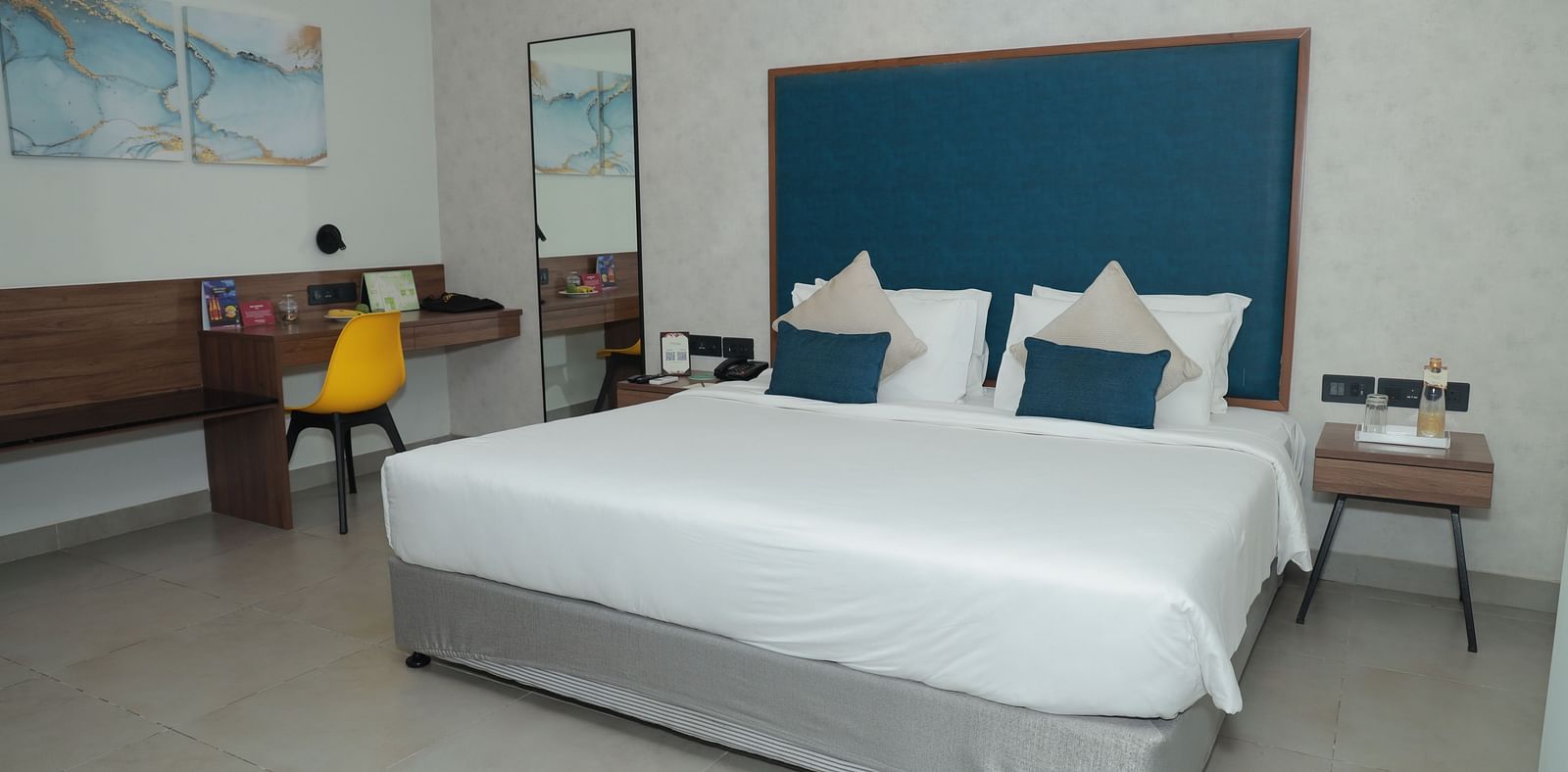 An image of a king size bed with a blue headboard in our suite in Tuticorin, with a work desk and chair on the right and a nightstand on the left - Regency Tuticorin by GRT Hotels