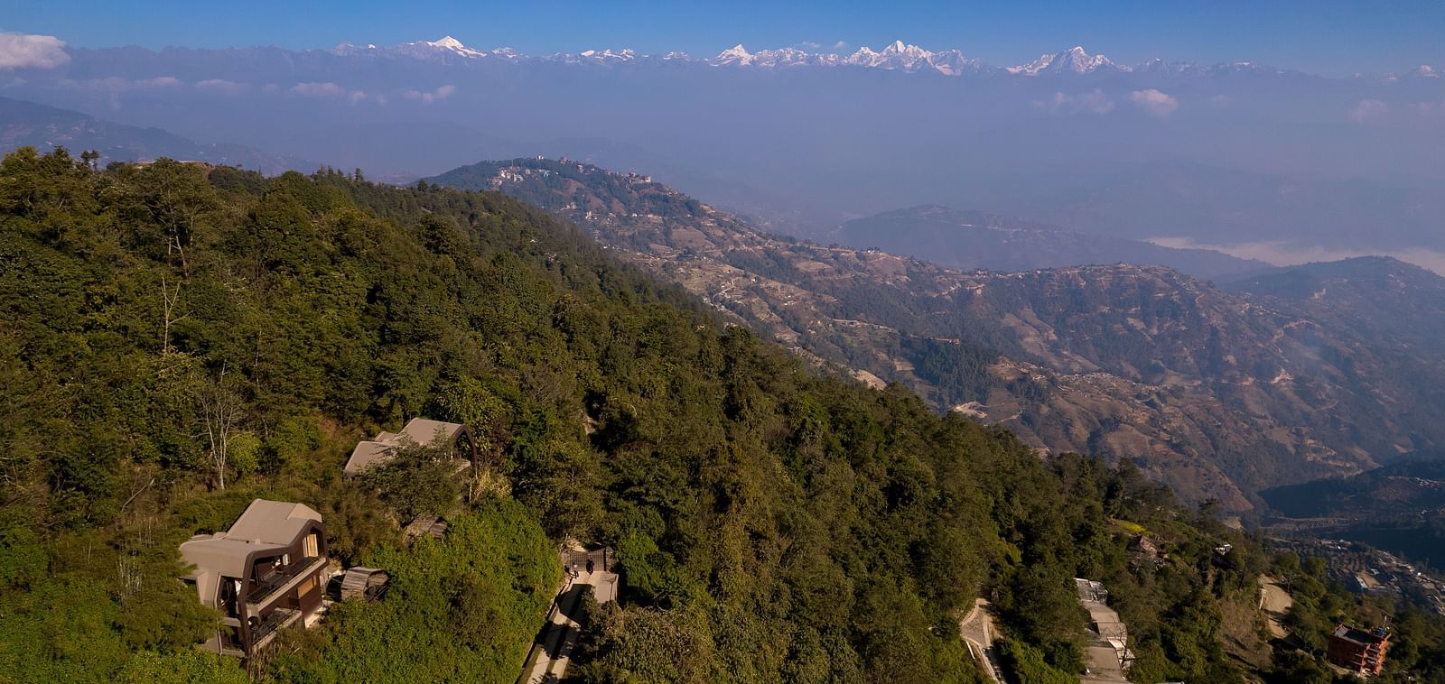 Aerial view of a hotel nestled in a lush green mountainside, with the distant snow-capped Himalayas in the background.