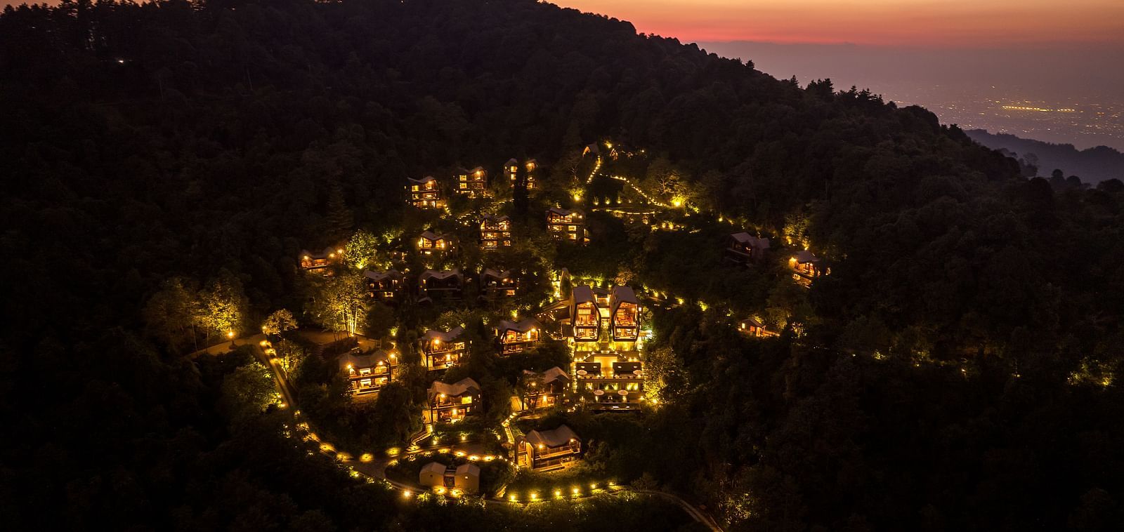 Nighttime aerial view of a resort illuminated with warm lights, nestled within a forested hillside.