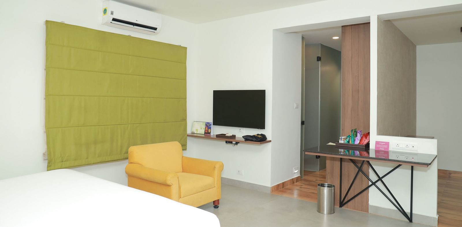 An image of the area in front of a bed in our suite, featuring a wall-mounted television, a window to the floor, a yellow chair, and a small table holding snacks - Regency Tuticorin by GRT Hotels