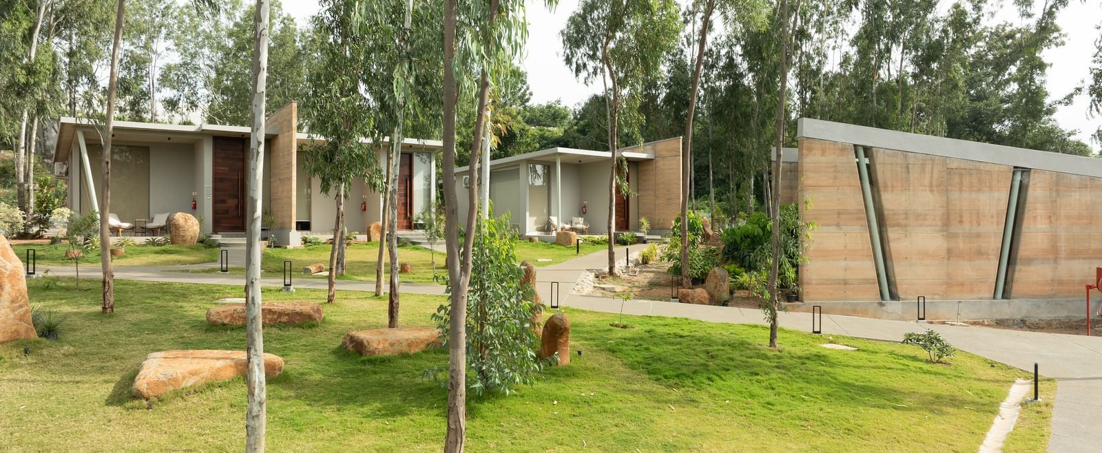 A wide exterior shot showing multiple modern residential units nestled within a landscaped area with trees and pathways | Gaia Studio | RD's Nature Retreat, Bangalore
