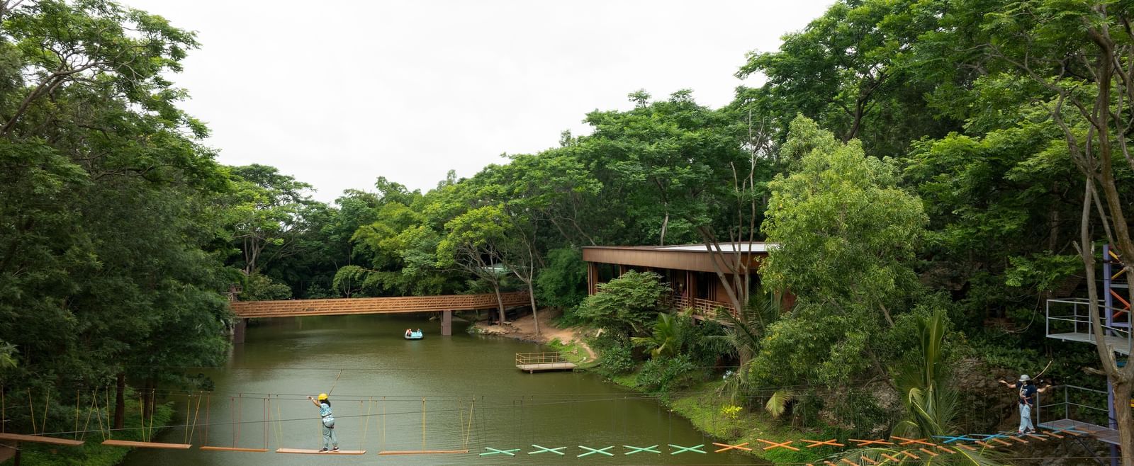 A calm body of water, possibly a lake or large pond, is framed by dense green trees and features a wooden structure extending over the water | RD's Nature Retreat, Bangalore