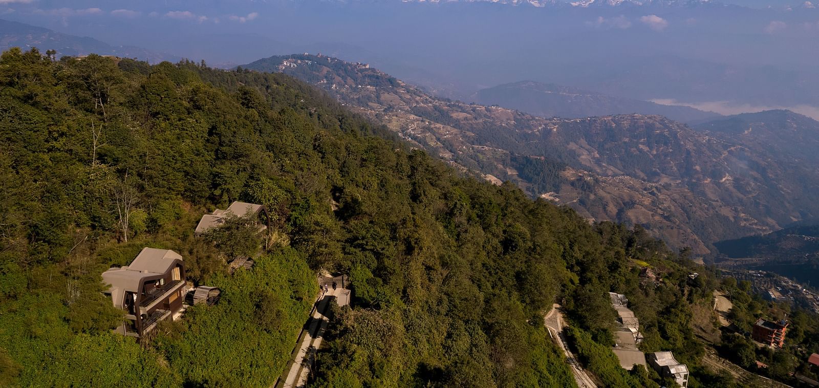 Aerial view of a hotel nestled in a lush green mountainside, with the distant snow-capped Himalayas in the background.