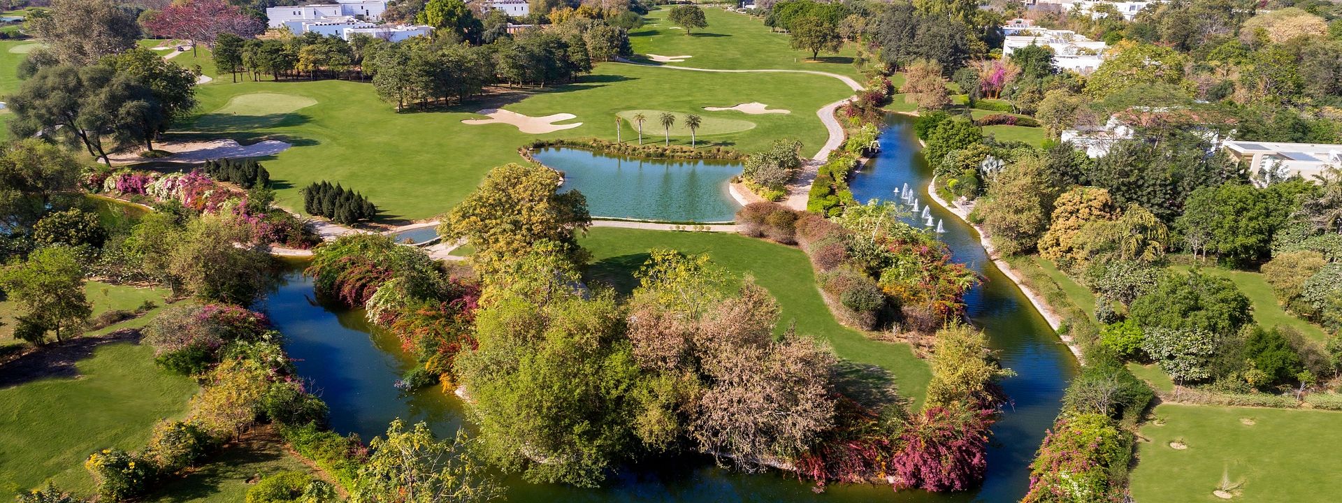 Aerial view of lush green golf course with winding water bodies and vibrant trees at Karma Chalets, with a city skyline in the background.