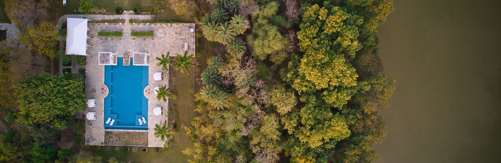 An aerial view of a rectangular pool adjacent to a building, surrounded by dense foliage and bordering a body of water- Shahpura Bagh.