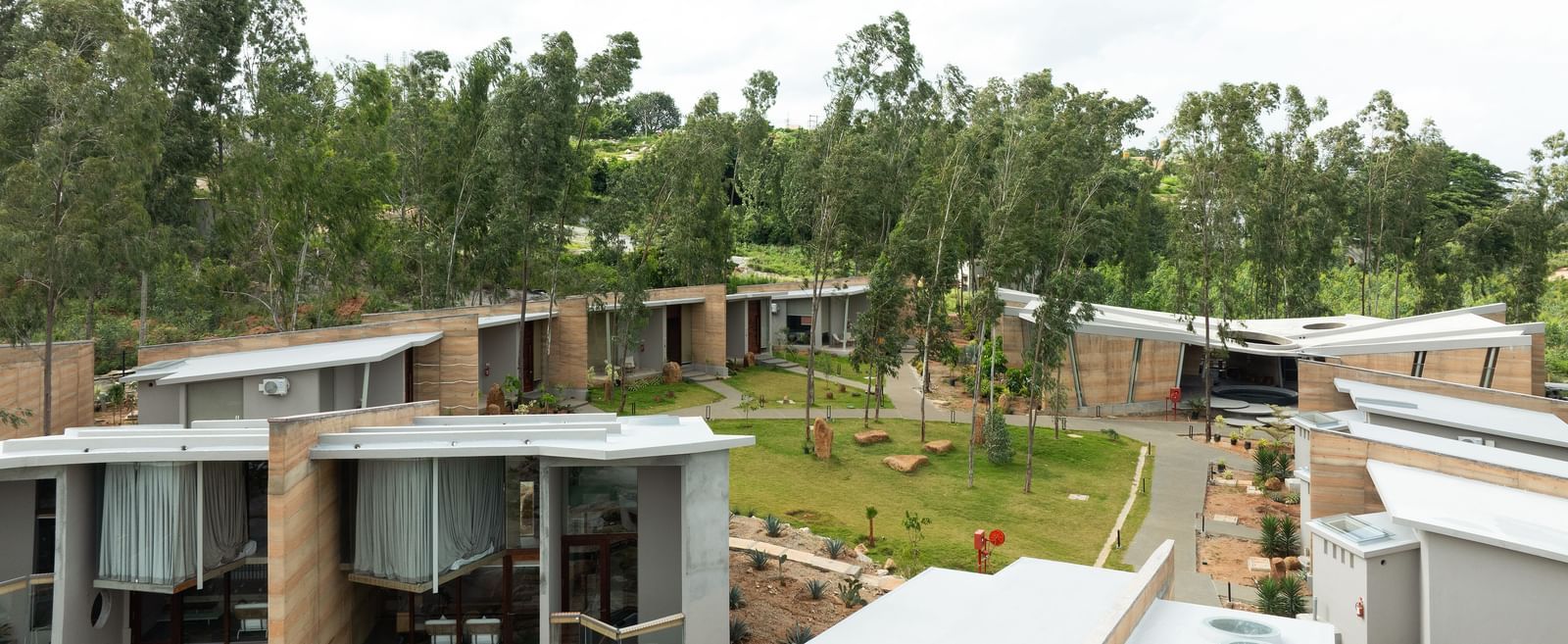 An aerial view of a residential complex with multiple buildings featuring diverse roof lines, surrounded by lush green foliage under a bright sky | Gaia Studio | RD's Nature Retreat, Bangalore