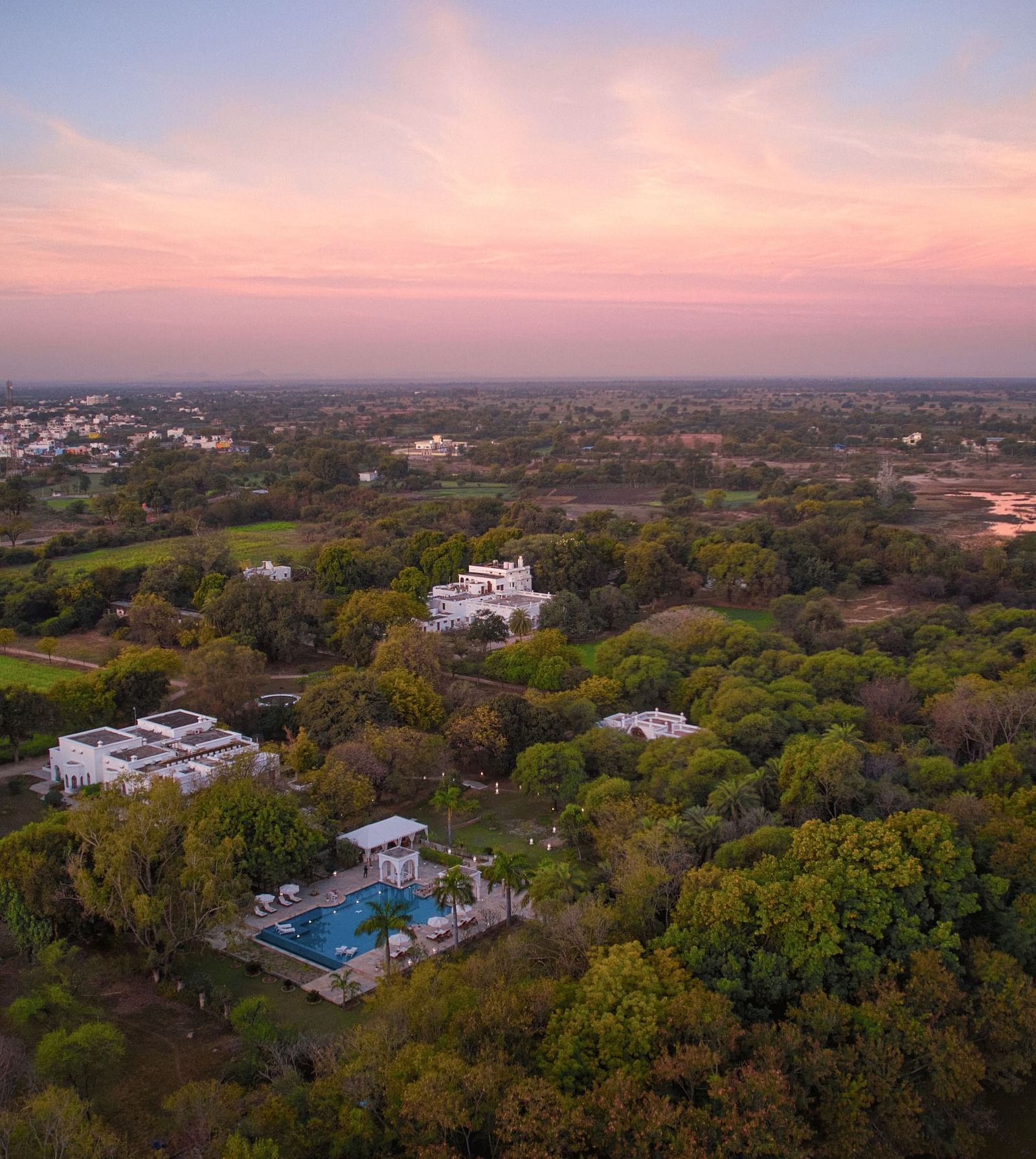 An aerial view of a landscape featuring structures nestled amidst dense foliage, bordering a body of water, under a sky transitioning at dusk- Shahpura Bagh.