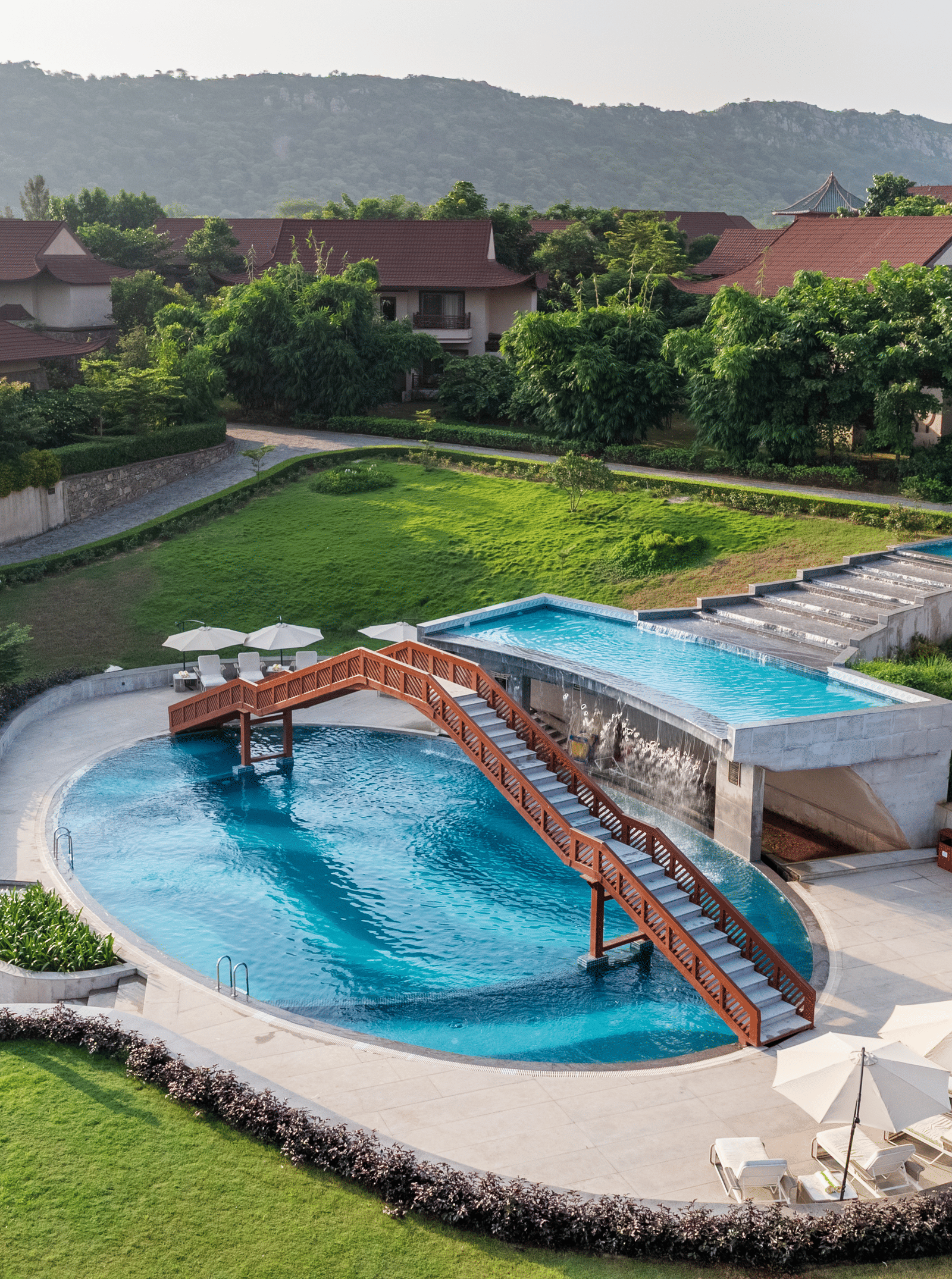 An aerial view of the swimming pool with an over-bridge going above it surrounded by nicely manicured lawns at Ananta Spa & Resort, Jaipur.