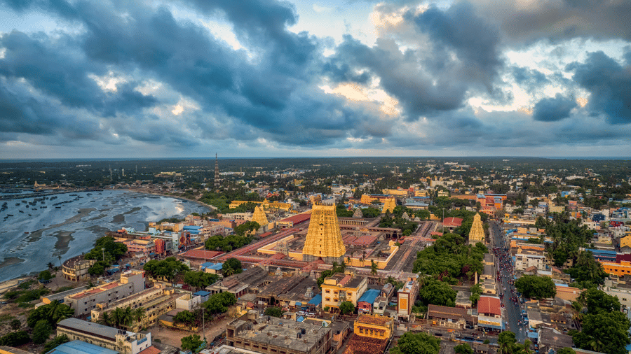 An aerial view of a densely populated city with a large Hindu temple tower (gopuram) dominating the skyline under dark clouds.