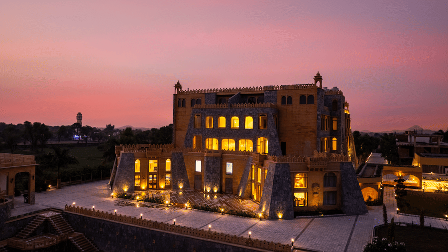 An evening view of EsthereaRaj Leela, Ranakpur, with lights shining from the windows against a vibrant pink and orange sunset.
