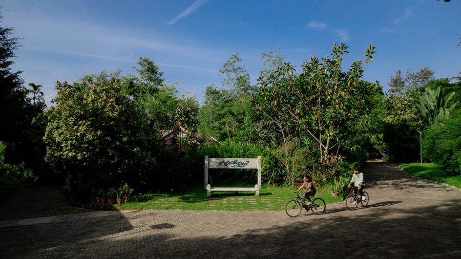 2 people riding bicycles along a dirt path past a wooden entrance sign board surrounded by green trees and bushes at Kadkani River Resort, Coorg.