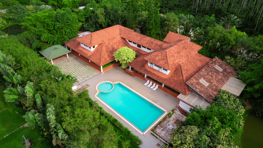 An aerial view of a multi-wing building with tiled roofs, a rectangular blue swimming pool, a smaller attached pool section, sun loungers, and surrounding trees at Kadkani River Resort, Coorg.