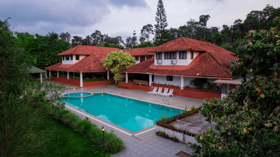 Outdoor swimming pool with blue water, sun loungers along the deck, and red tiled roof buildings in the background at Kadkani River Resort, Coorg.