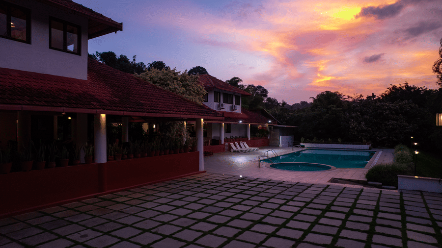 Paved outdoor courtyard next to a swimming pool and multi storey building under a cloudy sky at dusk at Kadkani River Resort, Coorg.