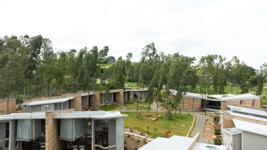 An aerial view of a residential complex with multiple buildings featuring diverse roof lines, surrounded by lush green foliage under a bright sky | Gaia Studio | RD's Nature Retreat, Bangalore