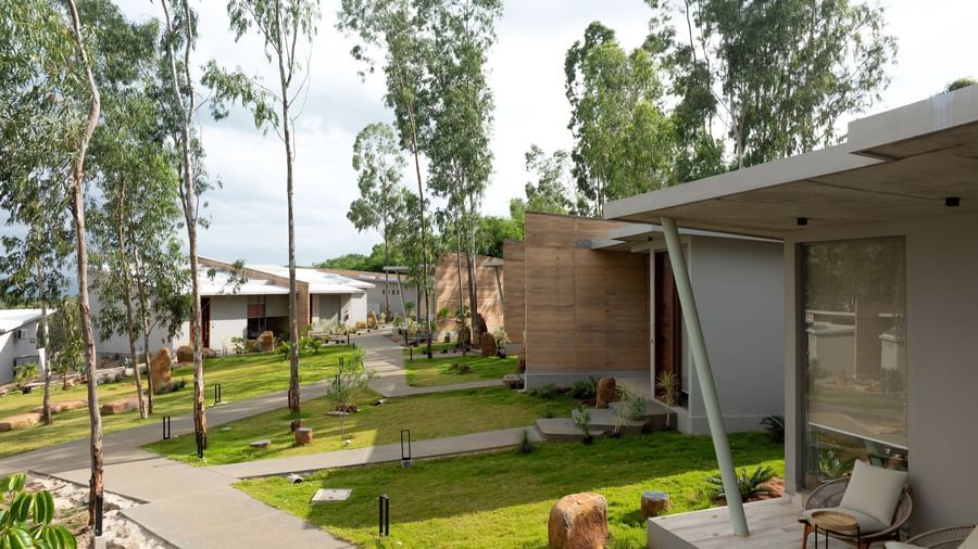 A ground-level view of contemporary buildings with sloped roofs and green lawns surrounded by trees | Gaia Studio | RD's Nature Retreat, Bangalore