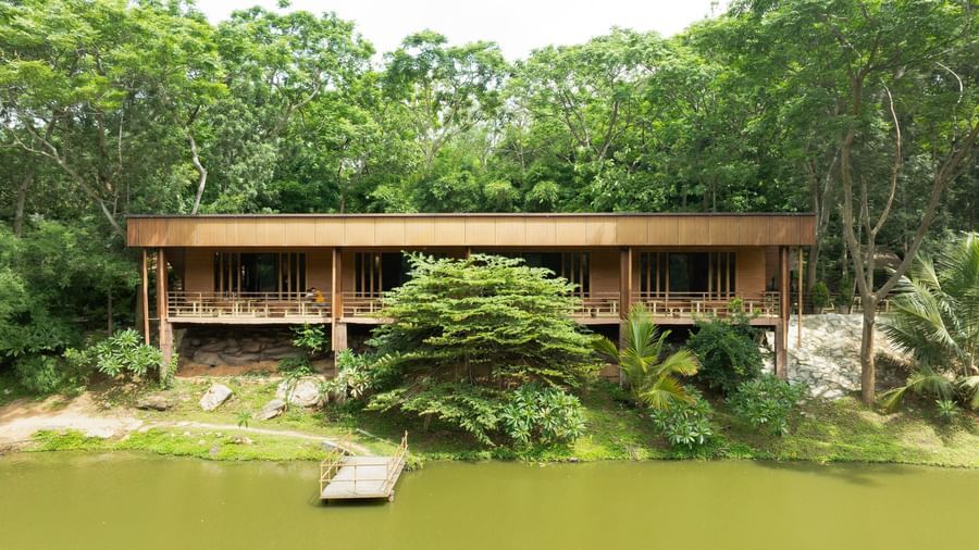 A modern building with a wooden exterior and a pier extends over a calm body of water, bordered by green trees | RD's Nature Retreat, Bangalore