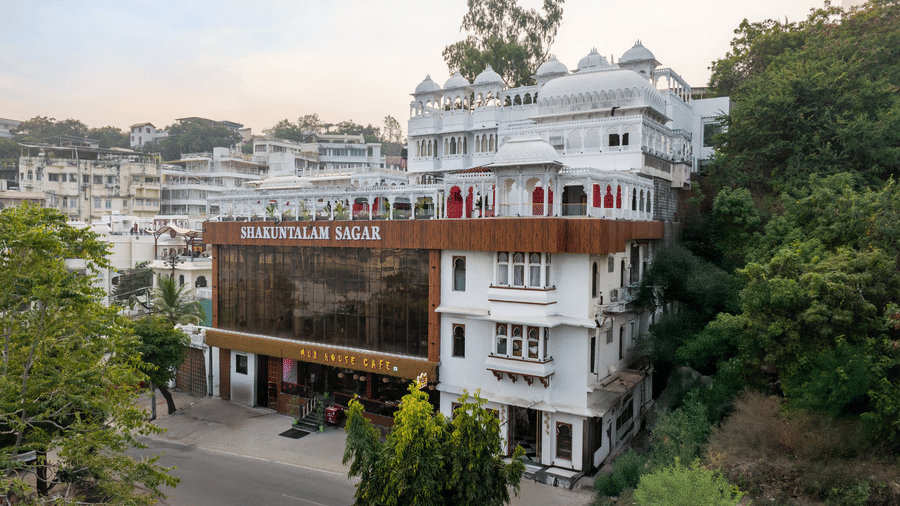 Wide exterior view of a grand white hotel building and its lower glass facade surrounded by foliage.