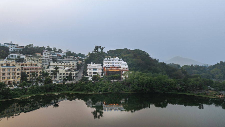 Wide exterior shot of a white hotel complex situated on a hillside overlooking a calm lake under a grey sky.