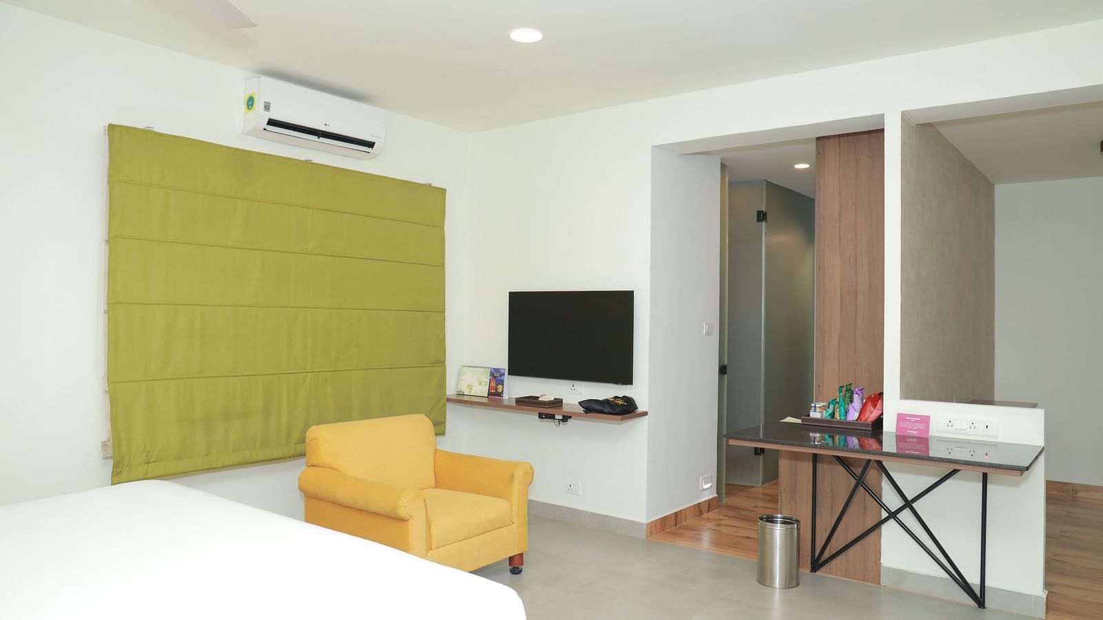 An image of the area in front of a bed in our suite, featuring a wall-mounted television, a window to the floor, a yellow chair, and a small table holding snacks - Regency Tuticorin by GRT Hotels