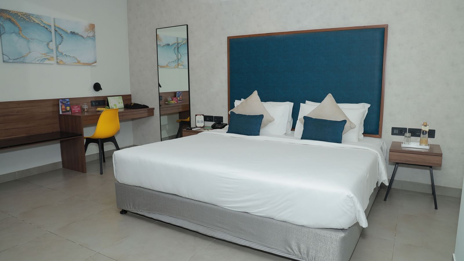 An image of a king size bed with a blue headboard in our suite in Tuticorin, with a work desk and chair on the right and a nightstand on the left - Regency Tuticorin by GRT Hotels
