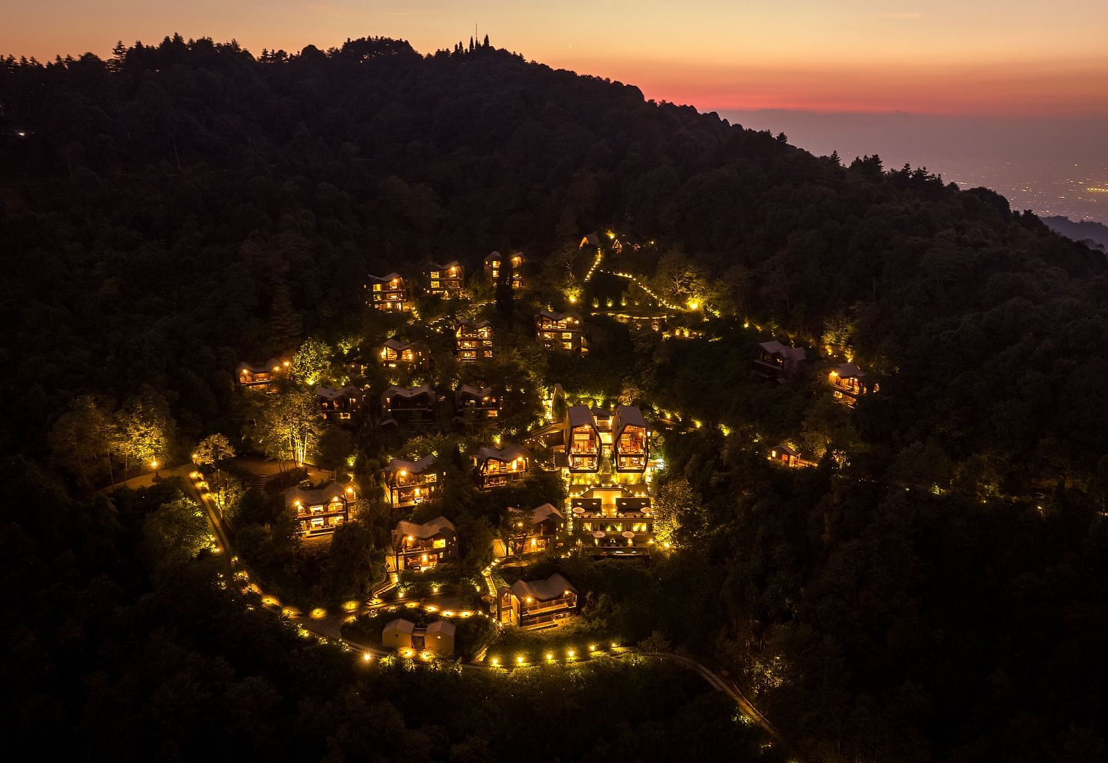 Nighttime aerial view of a resort illuminated with warm lights, nestled within a forested hillside.