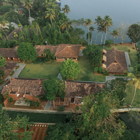 Aerial view of Kayaloram Ayurveda Retreat showing traditional tiled roof cottages surrounded by lush tropical greenery and palm trees on the edge of a calm backwater.