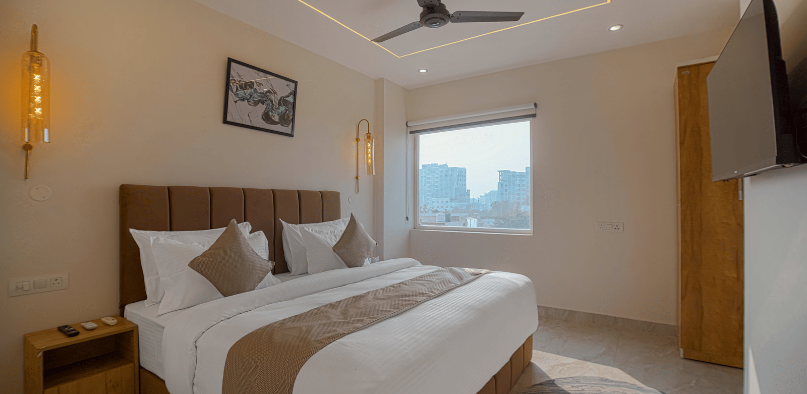 A contemporary room at Perfectstayz Premium Varanasi with a king size bed, beige and brown theme, a wall-mounted TV, and a window overlooking the city.