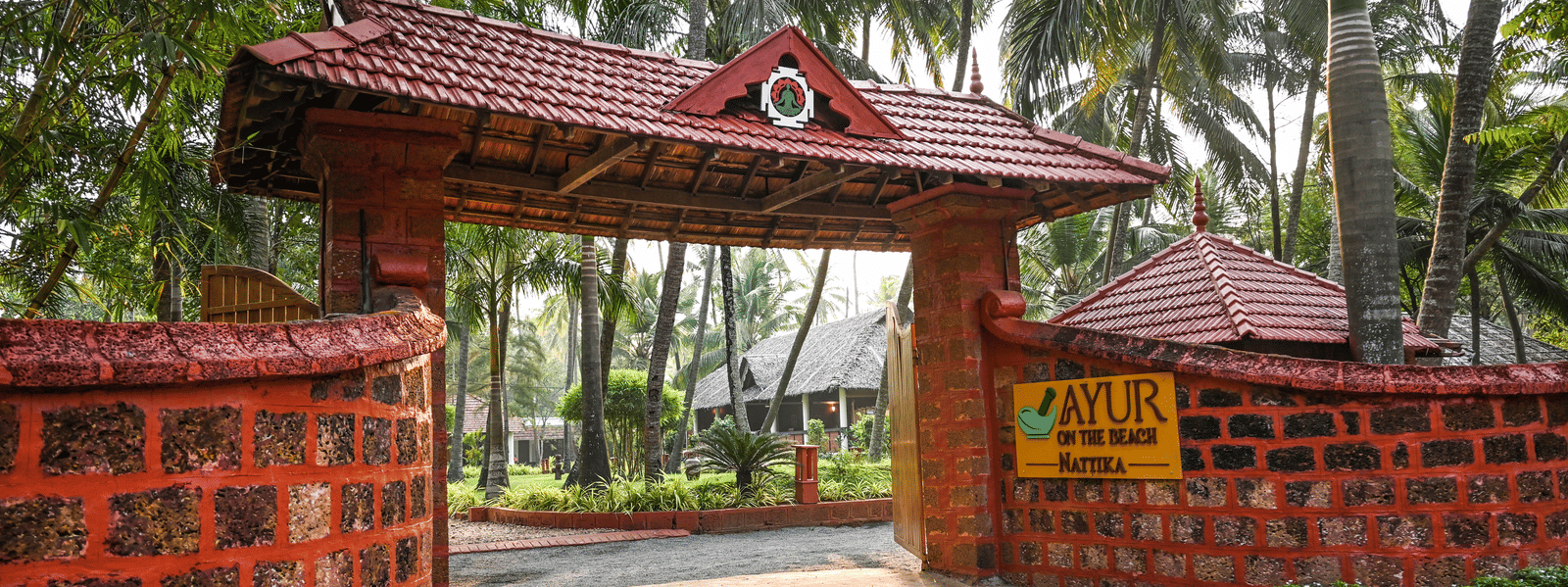 The ornate entrance gate of Ayur On The Beach Nattika topped with a traditional tiled roof and surrounded by greenery.