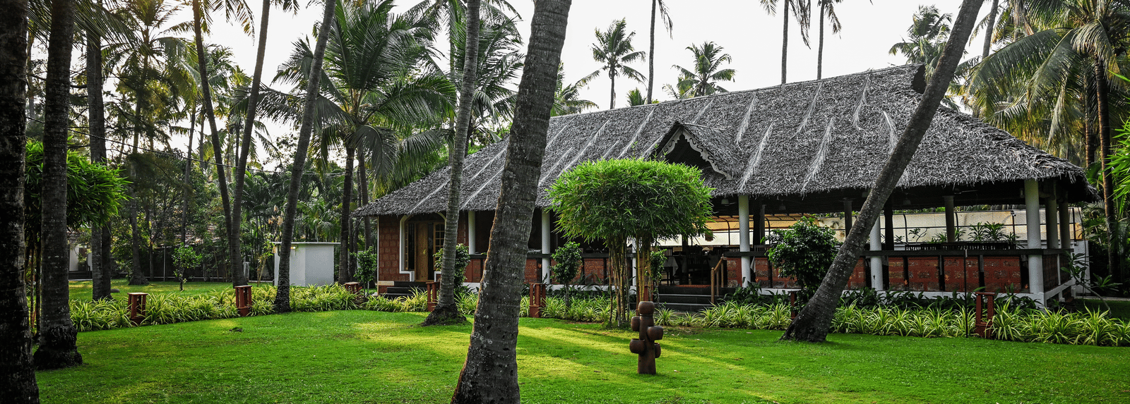 The lush, palm-shaded lawns and a traditional thatched-roof building at Ayur On The Beach Nattika during the golden hour.