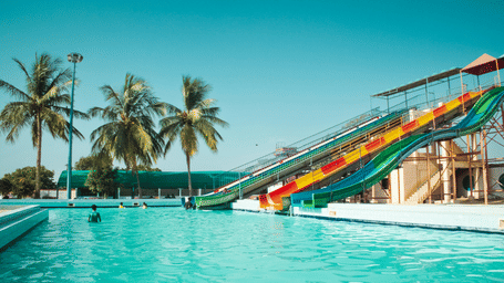 Outdoor resort swimming pool with a long water slide, palm trees, and clear blue water under a bright sky.