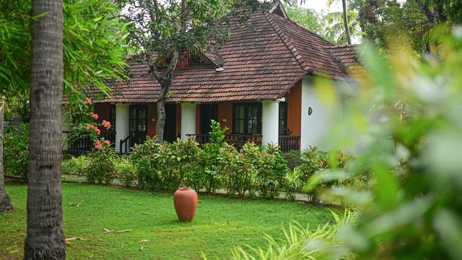 Daytime exterior view of Ayur Villas at Ayur on the Beach Nattika featuring a traditional tiled roof cottage surrounded by lush green lawns tropical plants and a clay pot in the foreground.