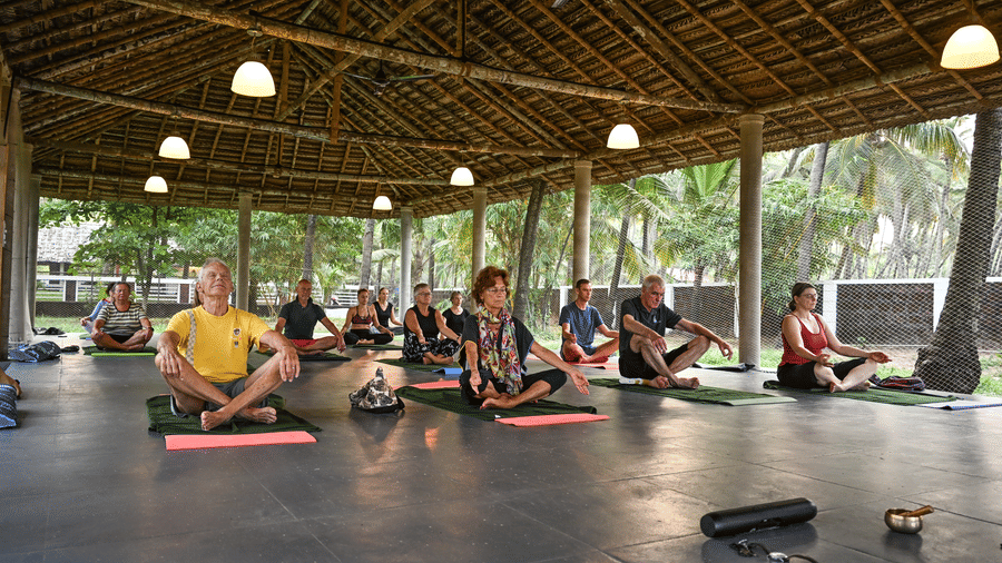 A group of guests seated on mats during a quiet meditation session under the large thatched roof pavilion at Ayur On The Beach Nattika.