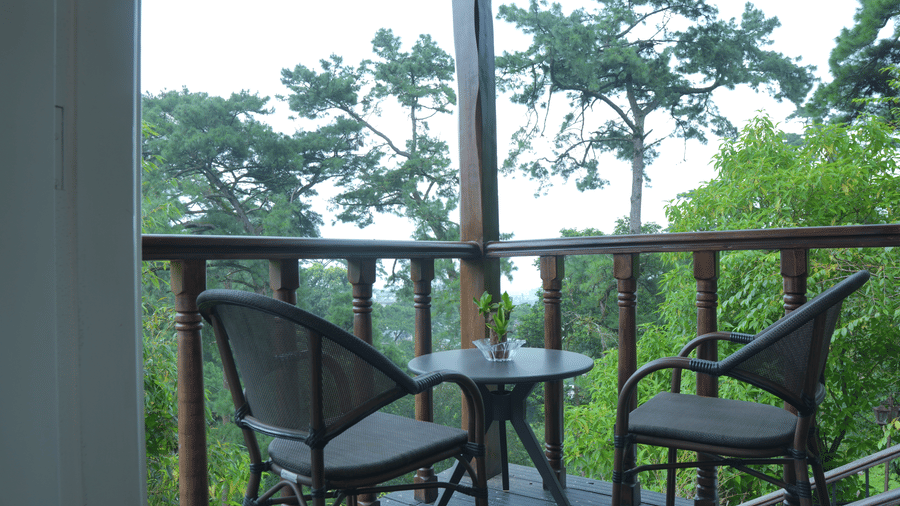 A balcony at The Heritage Club Tripura Castle Hotel and Spa, Shillong featuring 2 chairs, a small round table, wooden railings, and a view of trees.