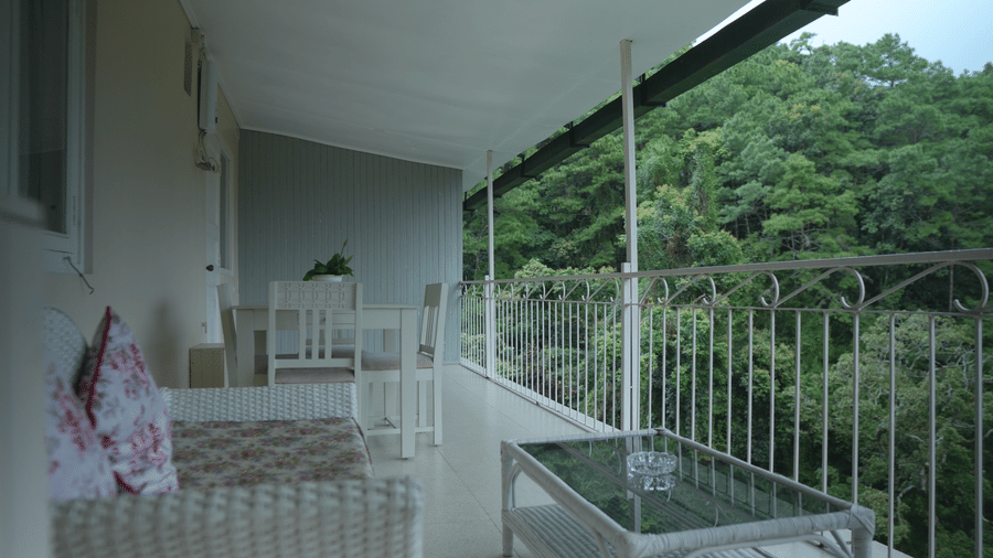 A balcony at The Heritage Club Tripura Castle Hotel and Spa, Shillong featuring a woven sofa, a coffee table, outdoor dining furniture, and views of forest foliage.  