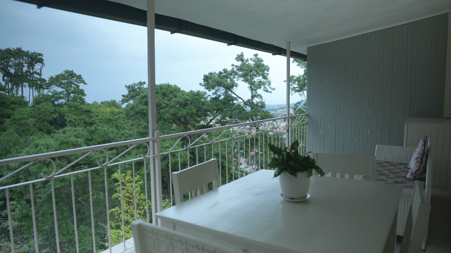A balcony at The Heritage Club Tripura Castle Hotel and Spa, Shillong featuring a white dining table with chairs, railings, and views of lush green trees.  