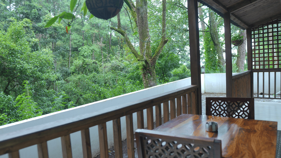 A outdoor balcony at The Heritage Club Tripura Castle Hotel and Spa, Shillong featuring a wooden dining table and chairs with views of the surrounding forest.  
