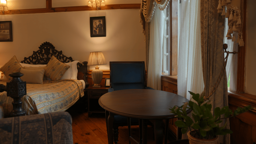 A bedroom at The Heritage Club Tripura Castle Hotel and Spa, Shillong featuring a king size bed, a dining table, 2 chairs, lamps, and curtains.  