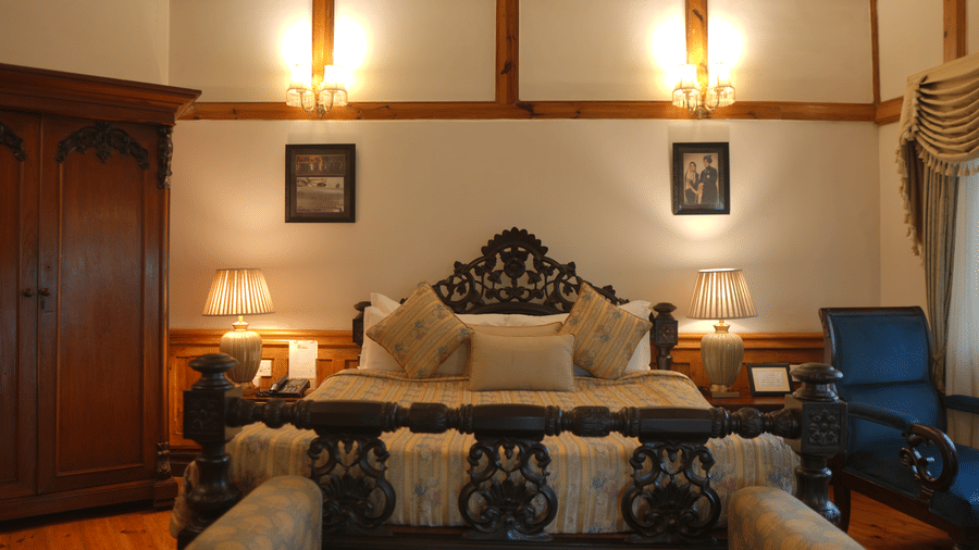 A bedroom at The Heritage Club Tripura Castle Hotel and Spa, Shillong featuring a king size bed with a carved wooden headboard, lamps, and a wardrobe.  