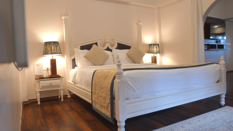 A bedroom at The Heritage Club Tripura Castle Hotel and Spa, Shillong featuring a four poster king size bed, bedside tables, lamps, and a doorway leading to another area.