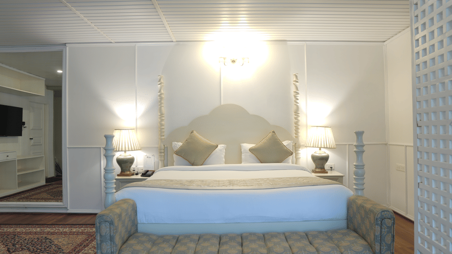 A bedroom at The Heritage Club Tripura Castle Hotel and Spa, Shillong featuring a king size bed, a bench, bedside tables, lamps, and a carpet on the floor.  
